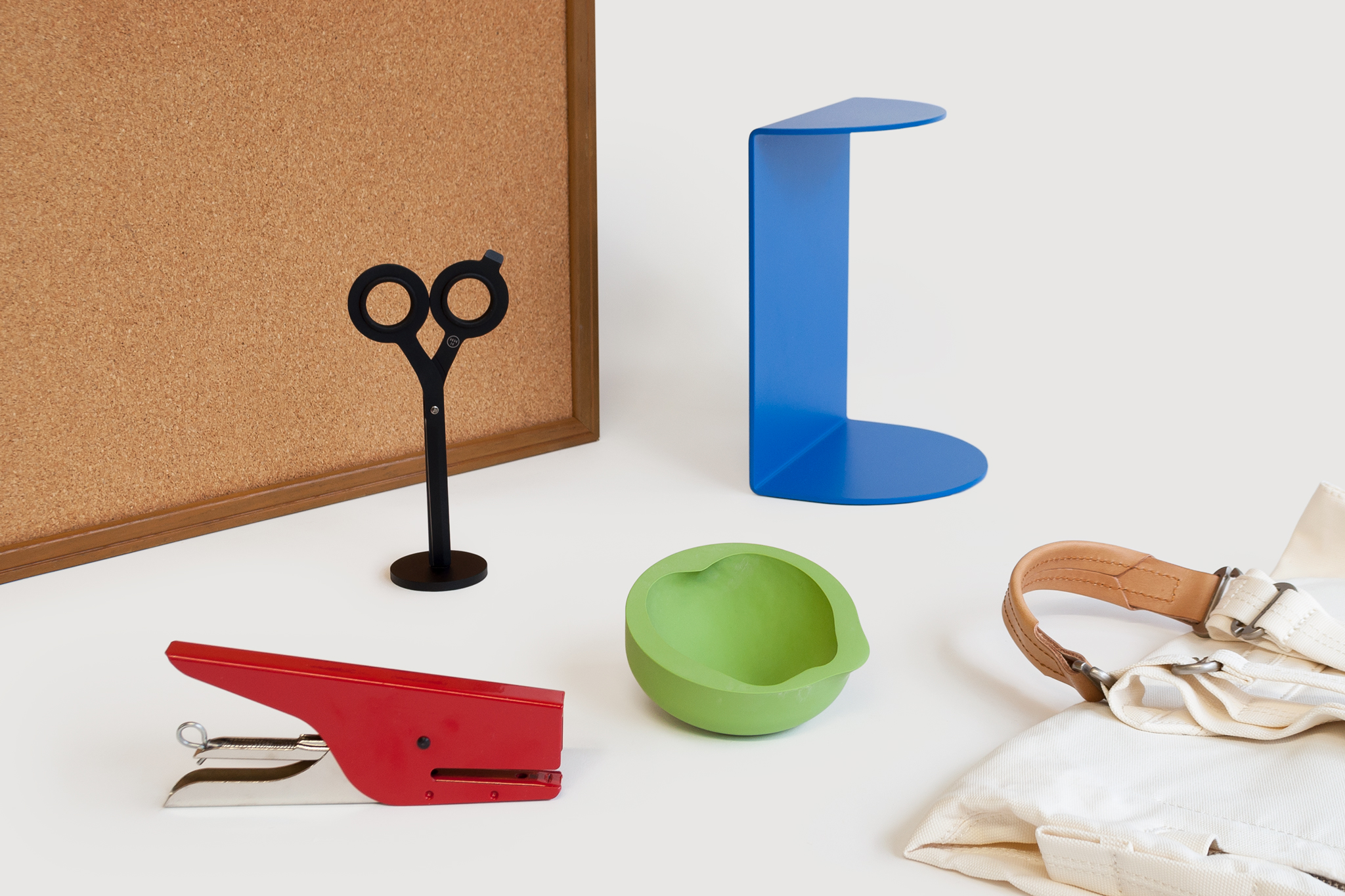 On a gray ground, a bulletin board stands upright behind a blue bookend, a red stapler, black standing scissors, a cream and brown leather backpack, and a green reverse molded apple vessel