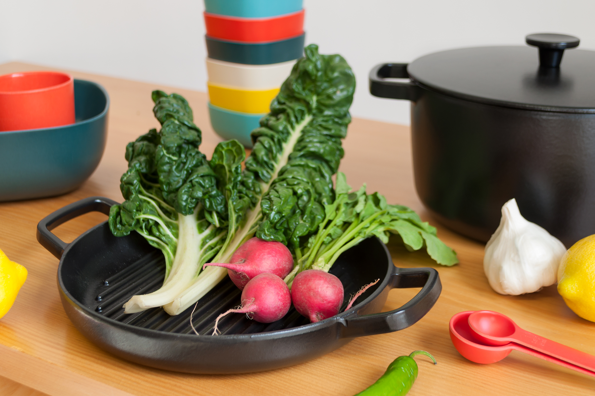 On a wooden surface, a black skillet holds fresh radishes and kale in the foreground, and is surrounded by colorful dishware, a black pot, and other ingredients