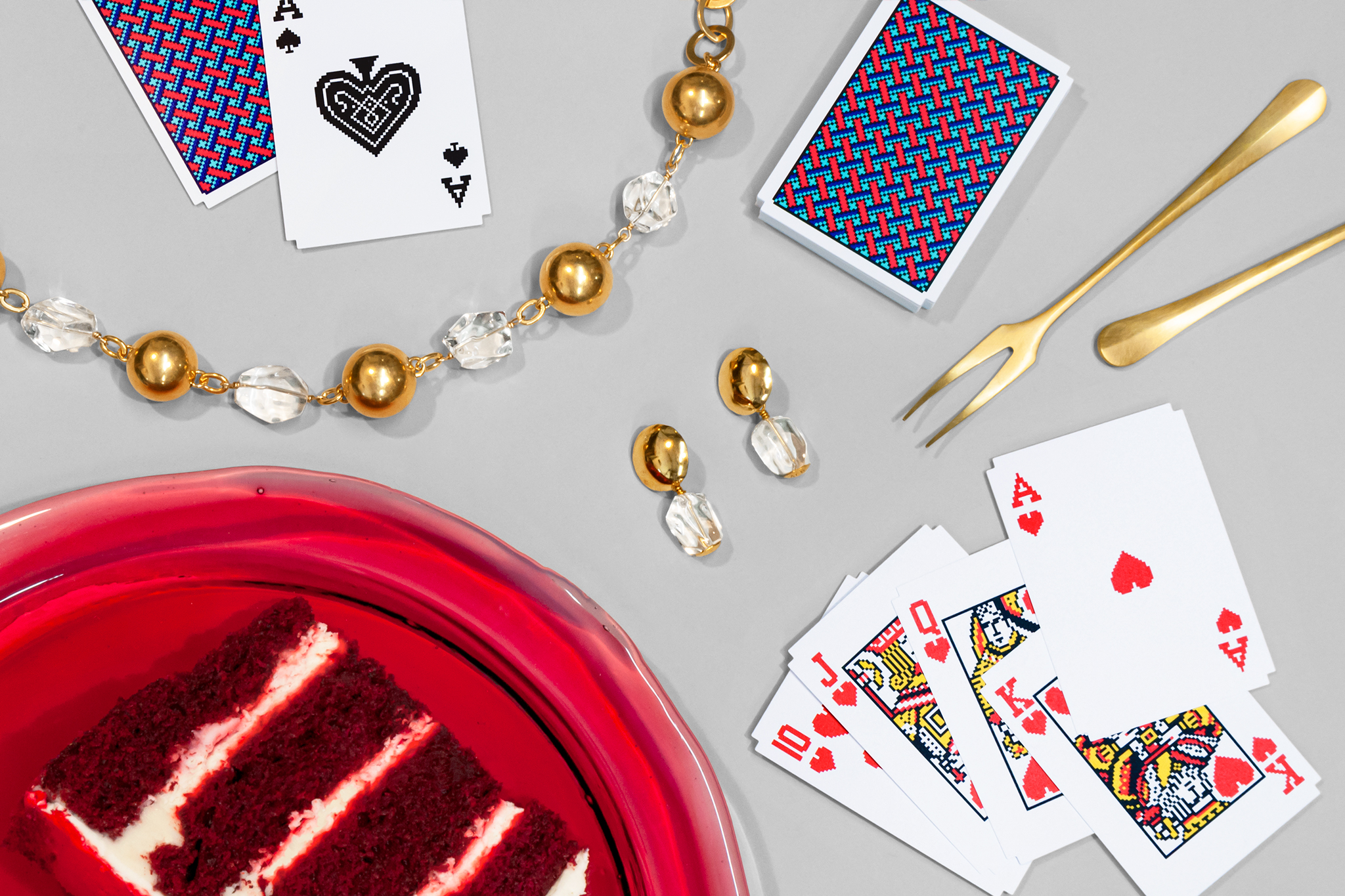 Scattered across a gray background: gold and quartz bauble jewelry, playing cards, brass serving utensils, and a resin tray with a slice of red velvet cake on it.
