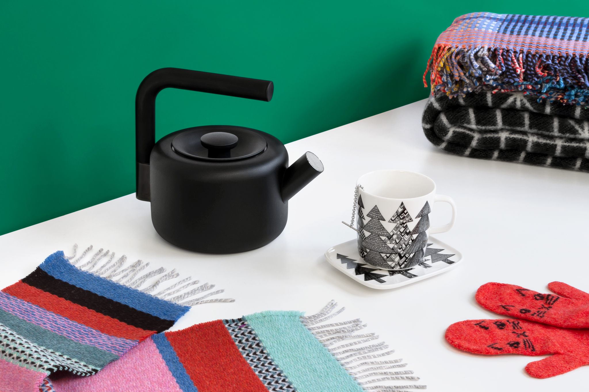 A modern black teapot faces a white mug illustrated with spruce trees on a matching dish. They are surrounded by a colorful striped scarf, orange mittens with cat faces, and a stack of blankets, all on a white tabletop with an emerald green wall behind.