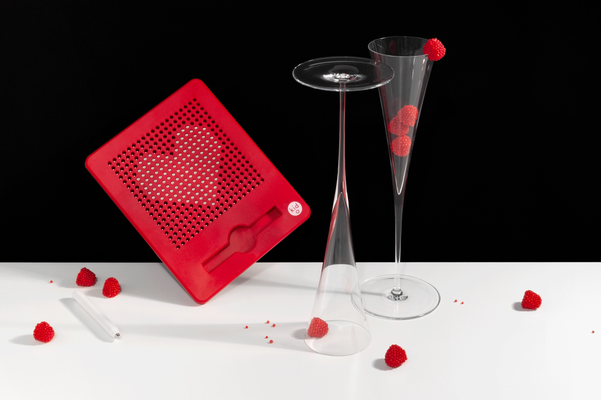 Two champagne glasses stand to the right of a red magnetic drawing board with a drawing of a heart that is animated to beat larger and smaller. There are red raspberry candies in and around the glasses, on a white tabletop against a black background.
